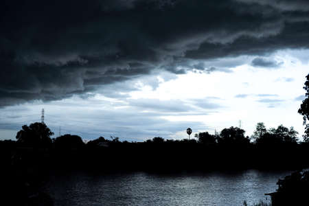 Silhouette of landscape of trees and electrical tower with rain cloud in the bright sky reflecting on the surface of the riverの写真素材