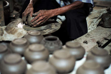 Selective focused on the dirty wrinkled skin hands of old man molding the clay work on the spinning wheel for making the jar with blurred group of clay jars in foregroundの写真素材