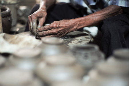 Selective focused on the dirty wrinkled skin hands of old man molding the clay work on the spinning wheel for making the jar with blurred group of clay jars in foregroundの写真素材