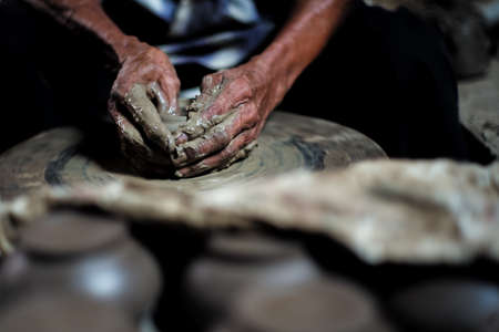 Selective focused on the dirty wrinkled skin hands of old man molding the clay work on the spinning wheel for making the jar with blurred group of clay jars in foregroundの写真素材