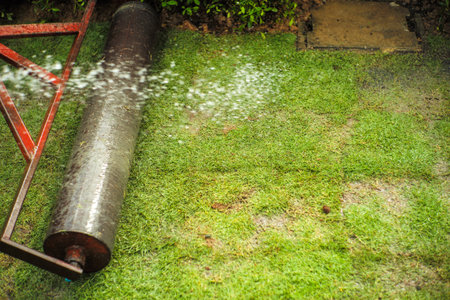 Selective focus on the grass field with motion blured of the heavy steel drum for pressing the lawn field during growing the grass and splashed watering in foregroundの写真素材