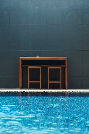 The wooden chairs and table in the modern design in front of the cement wall with blurred swimming pool in foregroundの写真素材
