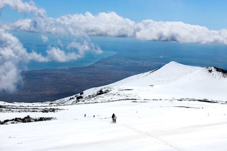 Amazing view from Mount Etna photographed with hikers going down on the snow and sea coast in the background. Magnificent clouds close to the top of the mountain. Etna, Sicily, Italy.の写真素材