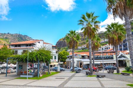 Ribeira Brava, Madeira, Portugal - Sep 9, 2019: Promenade of the Madeiran city photographed from the Atlantic ocean coast. People on the street. Outdoor restaurants, bars, and shops. Palm trees.のeditorial素材