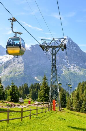 Yellow cable car in the Swiss Alps. Gondola going from Grindelwald to First in the Jungfrau region. Summer Alpine landscape with snowcapped mountains in the background. Transportation of tourists.の写真素材