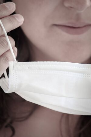 Detail photo of unrecognizable woman putting on a white medical face mask. Blurred background,...