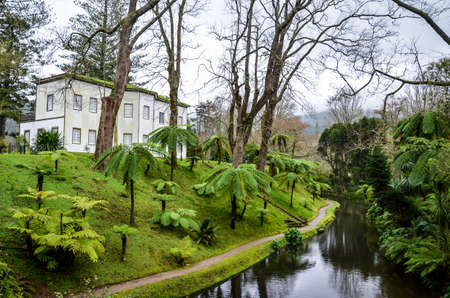 Furnas, Azores, Portugal - Jan 13, 2020: Botanical gardens in the Terra Nostra Garden area in Furnas. Water stream surrounded by green trees. House on the hill. Portuguese tourist destination.のeditorial素材