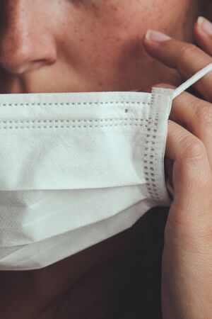Detail photo of unrecognizable white woman putting on a white medical face mask to prevent the flu. Blurred background, focus on the mask. Coronavirus, COVID-19 quarantine. Lockdown concept.の写真素材