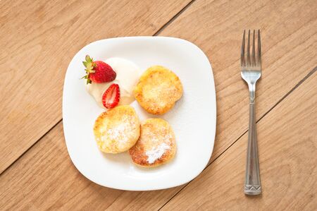 Breakfast of tasty round pancakes with cottage cheese with fragrant red ripe strawberries on a square white plate and coffee with milk cappuccinoの写真素材