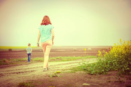 Young woman walking with son on rural road back to camera, vintage colorの写真素材