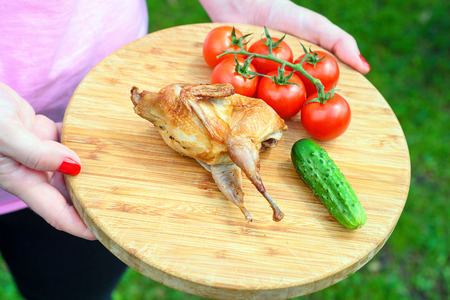 Delicious fried quail with fresh juicy vegetables on the wooden round board holding in hands. Prepared tomato, cucumber, partridge, quail. Roasted Partridge, quail grilling on sunny day. Picnic on sunny day. Culinary concept.の写真素材