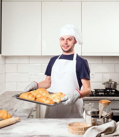 Happy baker showing tray with fresh croissant in the kitchen of the bakeryの写真素材