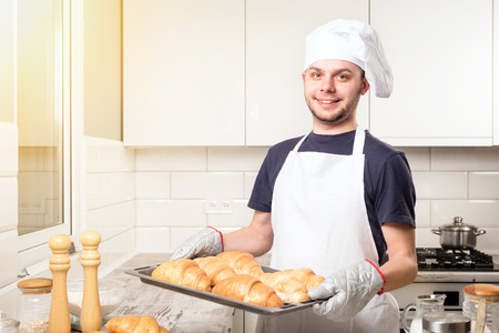 Happy baker showing tray with fresh croissant in the kitchen of the bakeryの写真素材