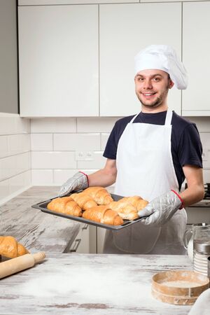 Happy baker showing tray with fresh croissant in the kitchen of the bakeryの写真素材