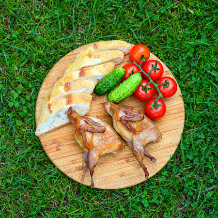 Delicious fried quail with fresh juicy vegetables on the wooden round board on green grass. Prepared tomato, cucumber, partridge, quail, slice of breads. Roasted Partridge, quail grilling on sunny day. Top view. Culinary concept with delicious food.の写真素材