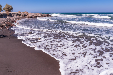 Waves hitting the black, volcanic sand of one of the beaches of Santorini island in Greece on a sunny, clear dayの写真素材