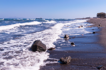 Waves hitting the black, volcanic sand of one of the beaches of Santorini island in Greece on a sunny, clear dayの写真素材