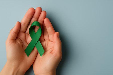A green ribbon held in hands symbolizes mental health awareness and support for emotional well being in calm and caring gesture on blue backgroundの素材