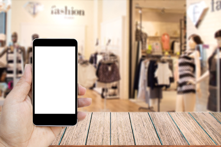 Mock up image of hand holding black mobile phone with blank white screen, Empty wood table top on blurred images shopping mall for background, perspective wood can be used for display products.の写真素材