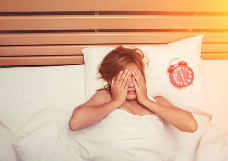 beautiful young woman take hands off her face on the bed in the morning when alarm clock is ringing don't want to wake upの写真素材