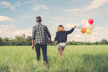 Back of happy young asian couple holding balloon and walk together on green grass.の写真素材