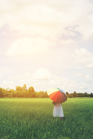 Back Of Young Beautiful woman holding multicolored umbrella in green grassland field and cloud blue sky.の写真素材
