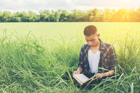 Young hipster man sitting on grassland reading book with nature around so happy.の写真素材