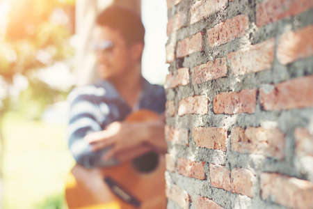 Handsome man leaning against a brick wall while playing the guitar.の写真素材