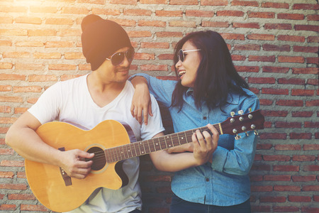 Hipster couple is hugging, looking away and smiling while standing outdoor playing guitar against brick wall, Dating spent great time together.の写真素材
