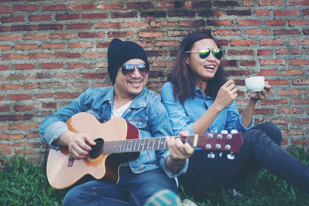 Hipster man playing guitar for his girlfriend outdoor against brick wall, enjoying together.の写真素材