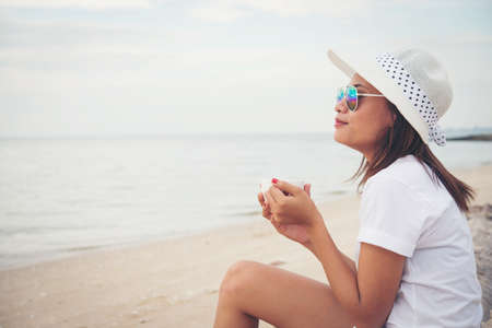 Beautiful young woman with coffee in hands sitting on the beach and watching the sunrise.の写真素材