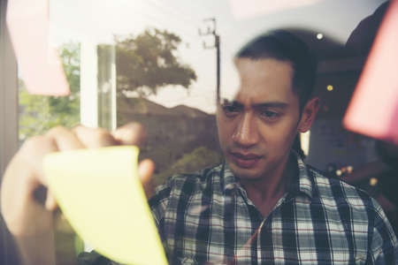 Young businessman reading sticky notes on the glass wall in office. Planning concept.の写真素材