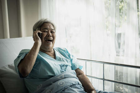 Female patient talking on phone while lying down on bed in hospital.の写真素材