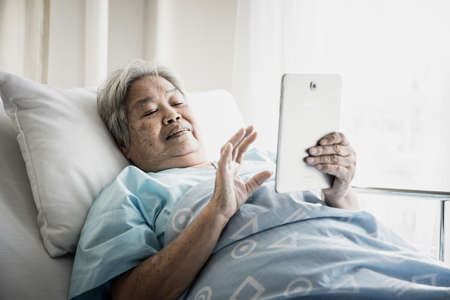 Old Female patient video call with tablet while lying down on bed in hospital.の写真素材