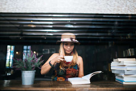 woman is holding a cup of hot cappuccino and reading a book, Business woman read a book in coffee shopの写真素材
