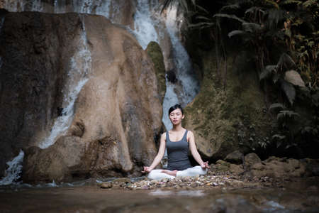 Young woman in yoga pose sitting near waterfallの写真素材