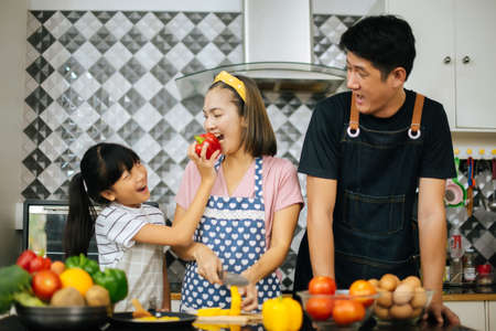 Cute girl help her parents are cutting vegetables and smiling while cooking together in kitchen at home.の写真素材
