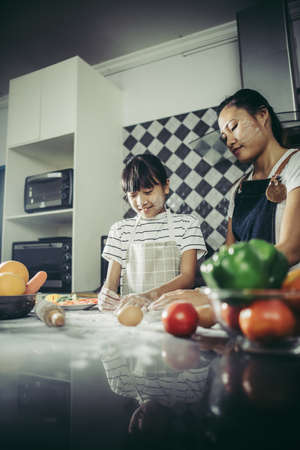 Cute little girl and her mom in aprons flattening the dough using a rolling pin in the kitchenの写真素材