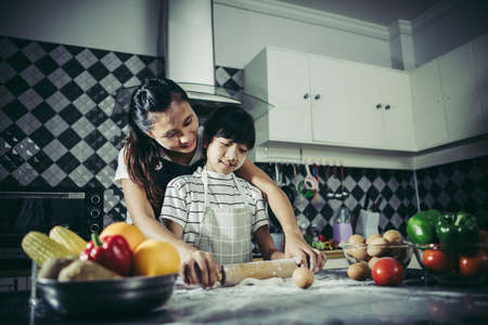 Cute little girl and her mom in aprons flattening the dough using a rolling pin in the kitchenの写真素材