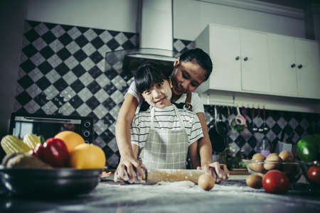 Cute little girl and her mom in aprons flattening the dough using a rolling pin in the kitchenの写真素材