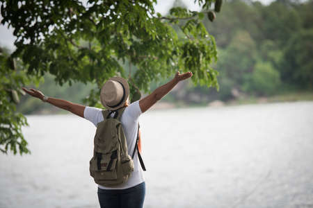 Young happy woman with backpack standing with raised hands and looking to the river. Woman traveling concept.の写真素材