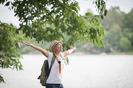 Young happy woman with backpack standing with raised hands and looking to the river. Woman traveling concept.の写真素材