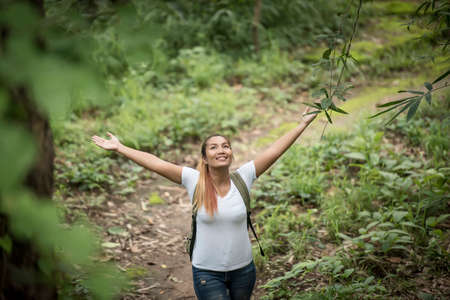 Young woman with backpack and raised up arms while walking through the forest.の写真素材