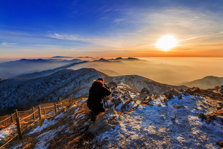 Professional photographer takes photos with camera on tripod on rocky peak at sunset.の写真素材
