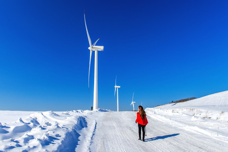 Beautiful girl walking in winter landscape of sky and winter road with snow and red dress and wind turbine.の写真素材