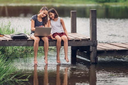 A 13-year-old sister and her 11-year-old sister sit Teach homework with a computer with an internet on a wooden bridge, The river behind the house happily.の写真素材