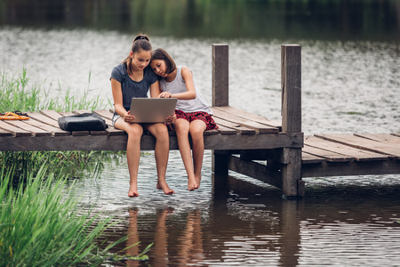 A 13-year-old sister and her 11-year-old sister sit Teach homework with a computer with an internet on a wooden bridge, The river behind the house happily.の写真素材