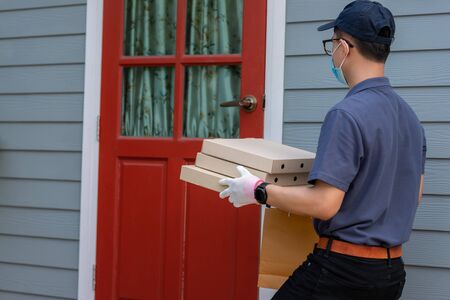 Asian delivery man employee in blue cap t-shirt uniform mask and medical gloves delivers takeaway food. Delivery service under quarantine, disease outbreak, coronavirus covid-19 pandemic conditions.の写真素材