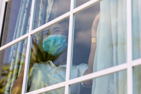 Coronavirus. Sick woman of corona virus looking through the window and wearing mask protection and recovery from the illness in home. Quarantine. Patient isolated to prevent infection. new normal.の写真素材