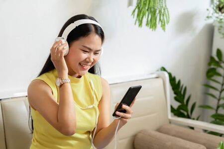 Asian smiling girl student wear wireless headphone study online with skype teacher in a coffee shop, woman learn language listen lecture watch at phone sit in cafe, distant education, New normalの写真素材